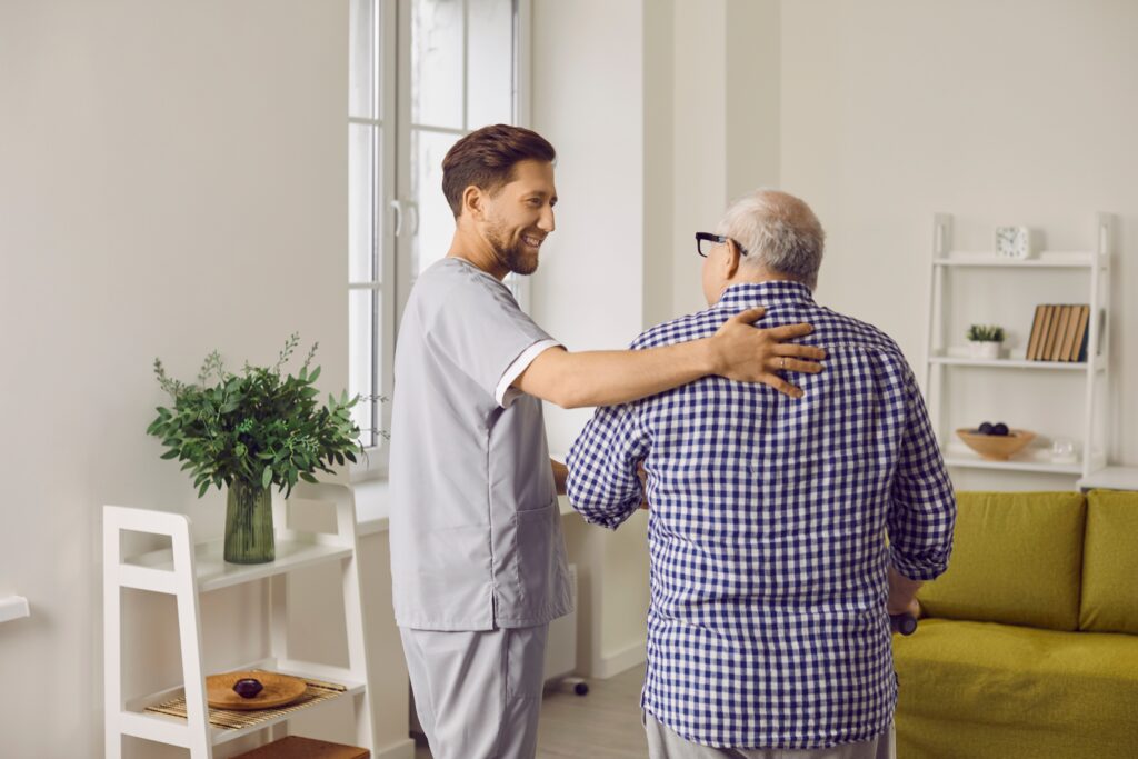 Smiling Young Male Caregiver In Uniform, Hugging Elderly Man During