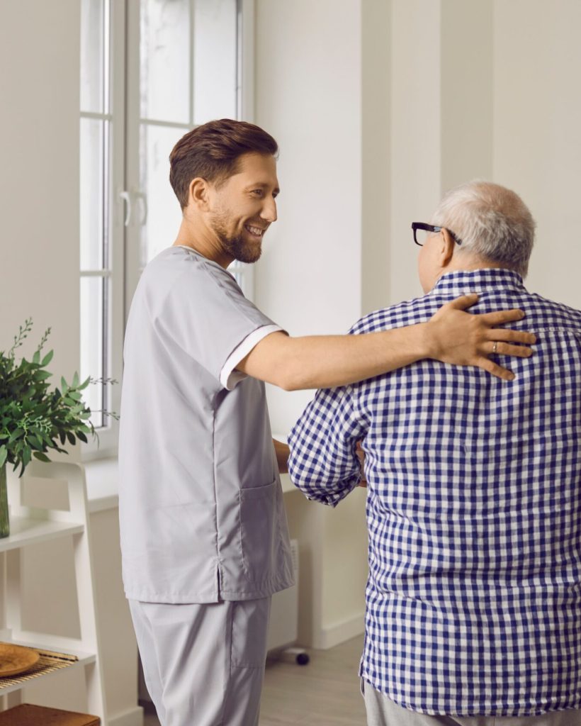 Smiling Young Male Caregiver In Uniform, Hugging Elderly Man During